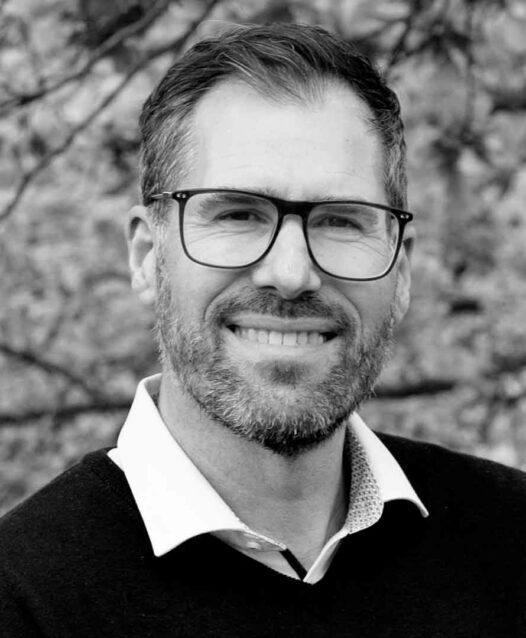 En man med glasögon ler mot kameran, med trädgrenar i bakgrunden.