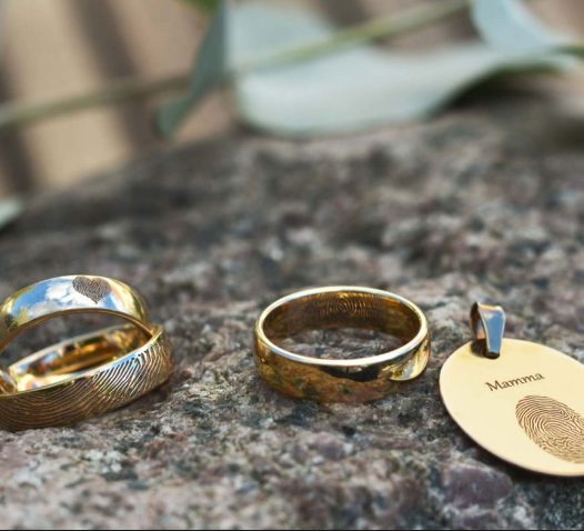 Guldringar och ett guldhänge med fingeravtryck ligger på en sten. Hänget har texten "Mamma" ingraverad.