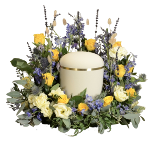 En vit urna med ett gulddekorerat band omgiven av en blomsterkrans med gula rosor, vita blommor och blå blommor.